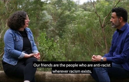 Two people sitting on a log in the bush with the words on screen "Our friends are those who are anti-racist, wherever they are"