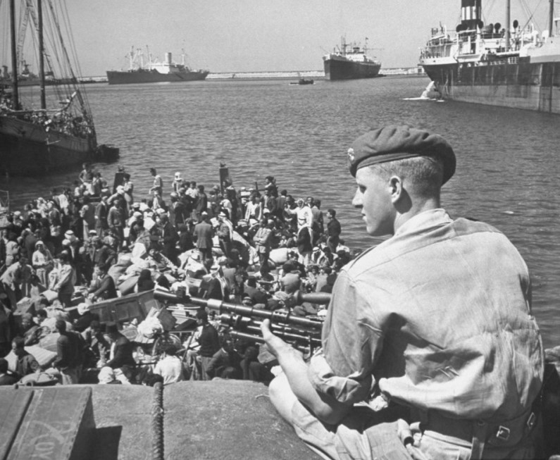 Historical photo of man in uniform with gun, sitting over boatloads of people