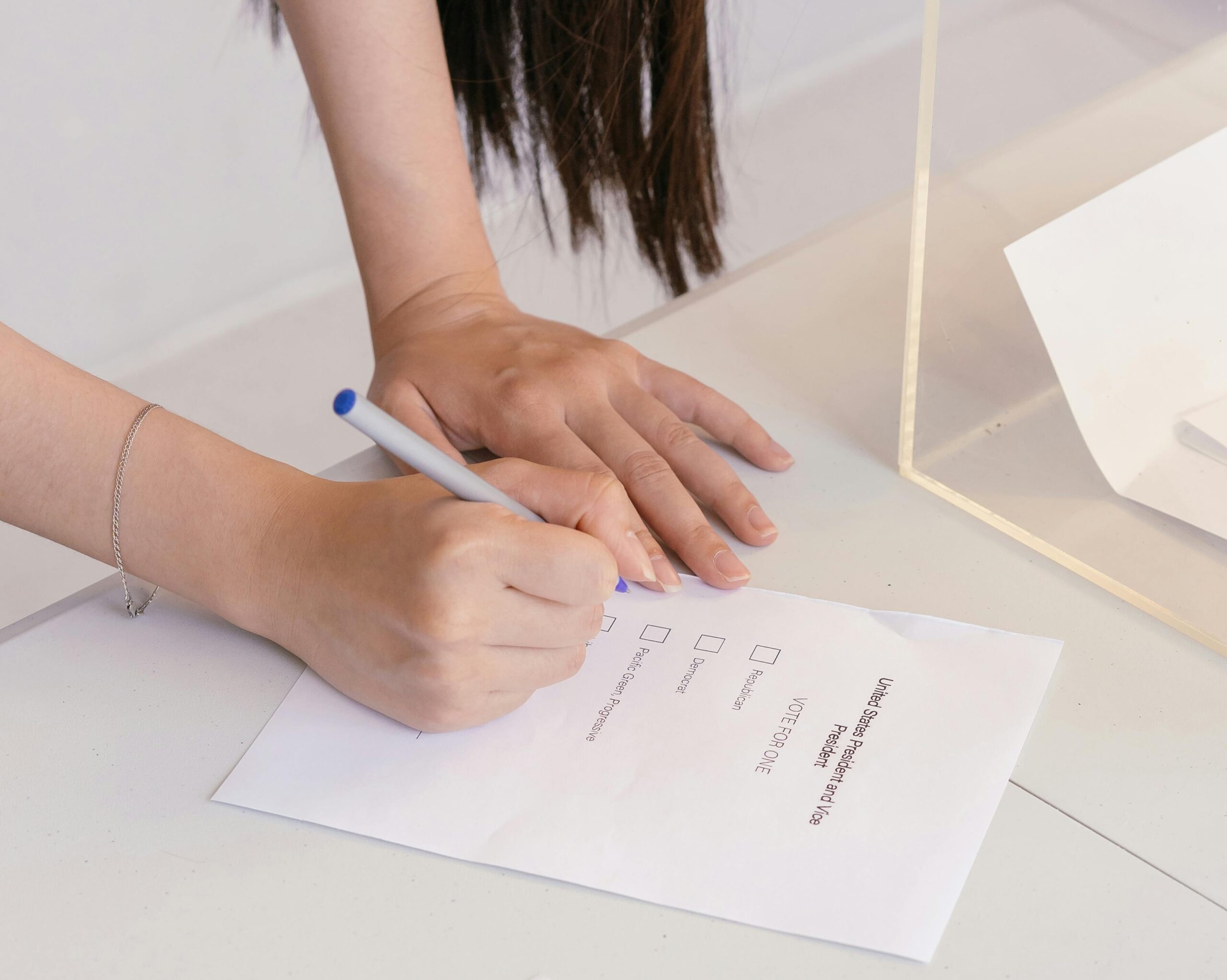 A person with long dark hair bending over a white table, holding a pen and filling out a ballot paper.